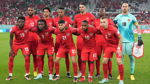 Red-uniformed soccer team poses for a group photo on the field before kickoff, stadium crowd behind.