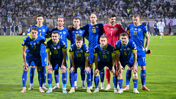 Bosnia-Herzegovina and opponent soccer teams pose on the field before kickoff, players in blue and red kits.