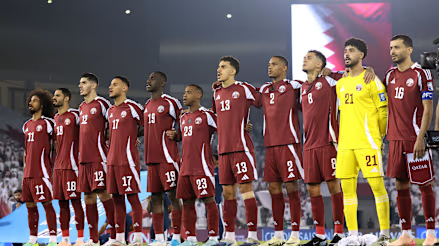 Football team lineup in maroon kits and goalkeeper in yellow, standing on stadium pitch before kickoff. Qatar.