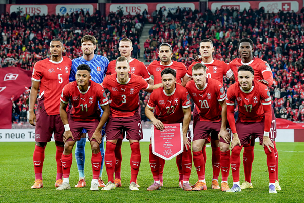 Football team lineup in maroon kits and goalkeeper in yellow, standing on stadium pitch before kickoff. Qatar.