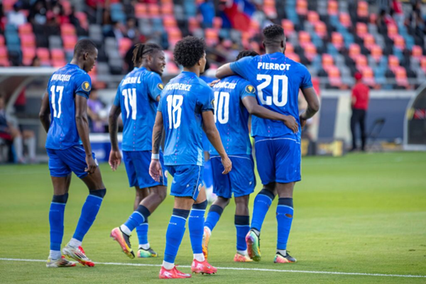 Soccer players in blue kits huddle on the field during a match, with “PIERROT 20”visible Haiti.