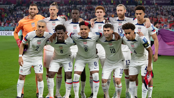 US soccer team posing together on field in white uniforms before a match, stadium crowd behind them.