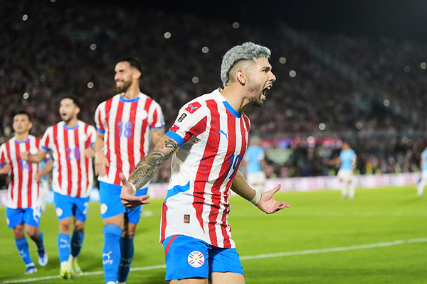 Striker in red-and-white striped kit celebrates on stadium pitch while teammates trail behind. Paraguay