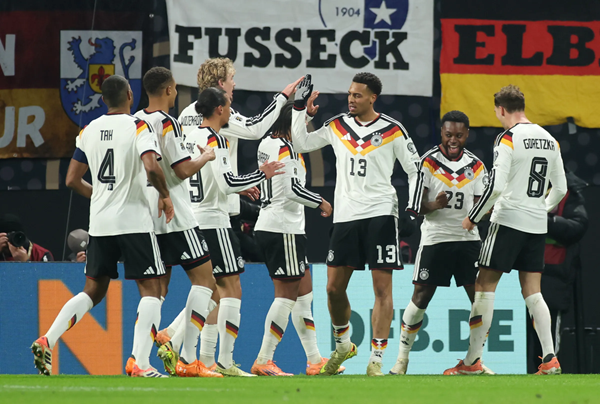 German soccer players in white celebrate together on the field, with banners reading FUSSECK and ELB.