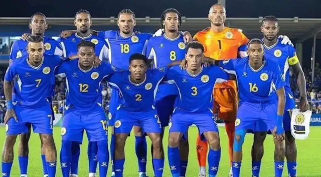 Curacao Soccer team posing in blue uniforms with a goalkeeper in orange, standing on a stadium field.