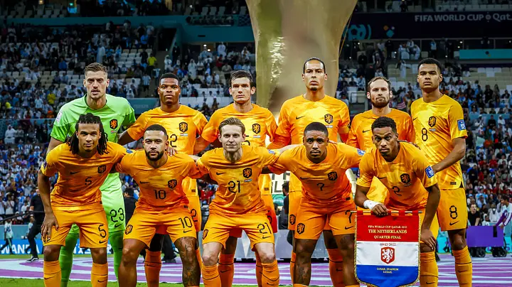 Netherlands national football team lineup posing in orange kits before a stadium crowd, with trophy behind.