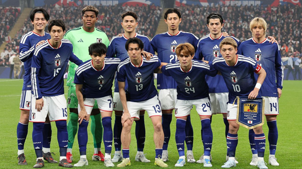 Japanese national soccer team posing together on a stadium field in blue uniforms, holding a trophy plaque.