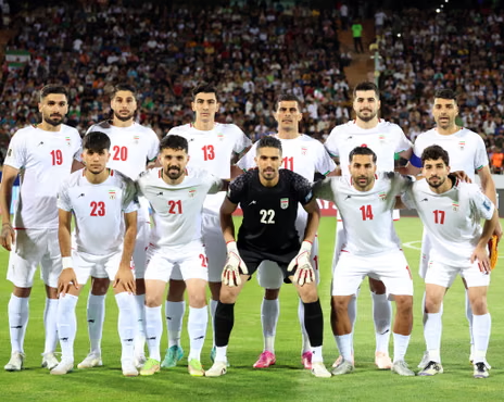 Soccer team lineup posing on field in white uniforms with goalkeeper in black, stadium crowd behind. Iran.