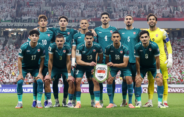 Football team in teal kits posing for a stadium photo, goalkeeper in yellow, fans filling the background. Iraq
