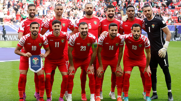 Soccer team lineup posing on field in red uniforms with goalkeeper in black, stadium crowd behind. Jordan.
