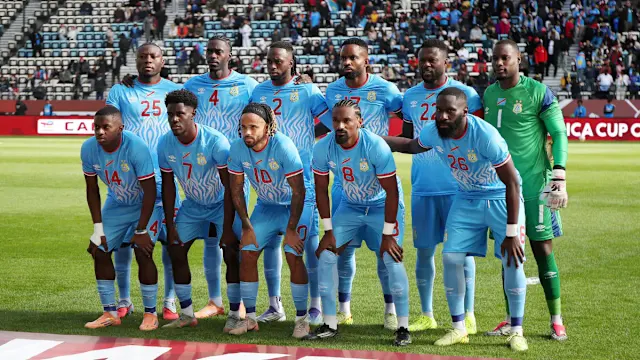 Soccer team in light blue uniforms posing on the field before a match, with goalkeeper in green. Congo Dr.