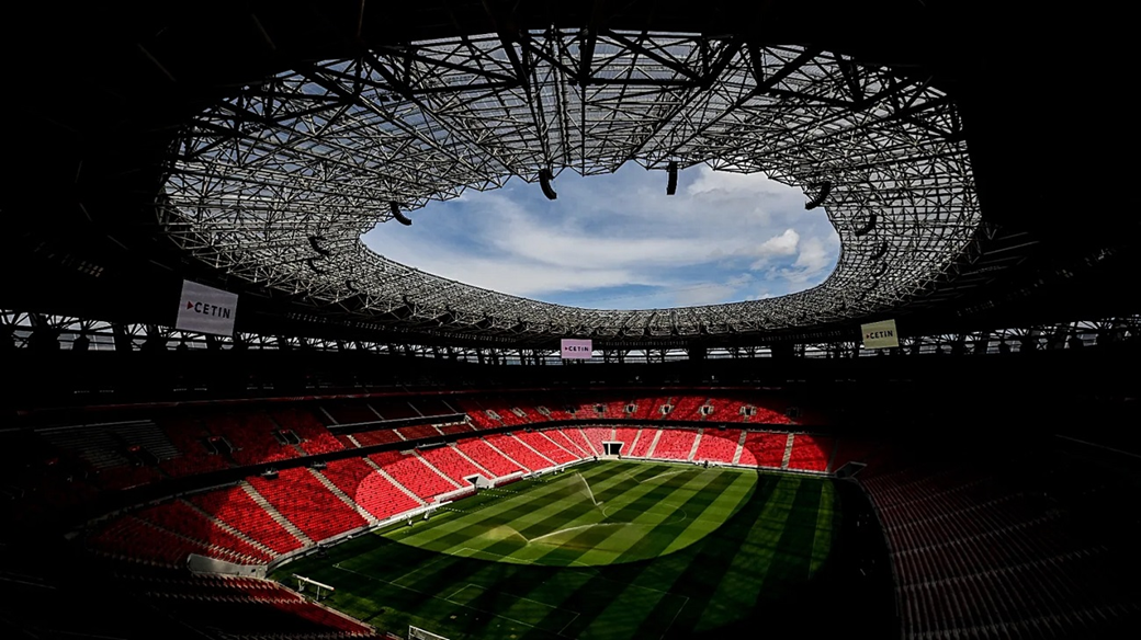 View of a football stadium interior with green pitch, red seats, and a curved roof opening to sky.